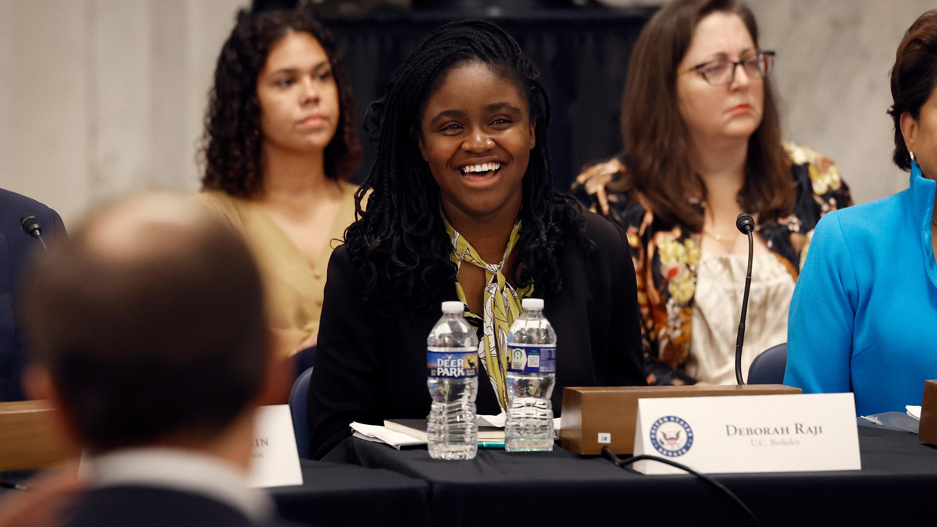 a woman sitting at a table smiling with people sitting behind, beside and in front of her